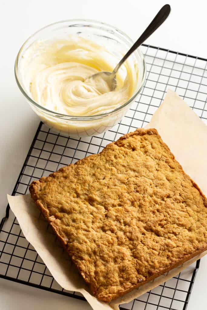 A bowl of cream cheese frosting and carrot cake slice sit on a wire baking rack.