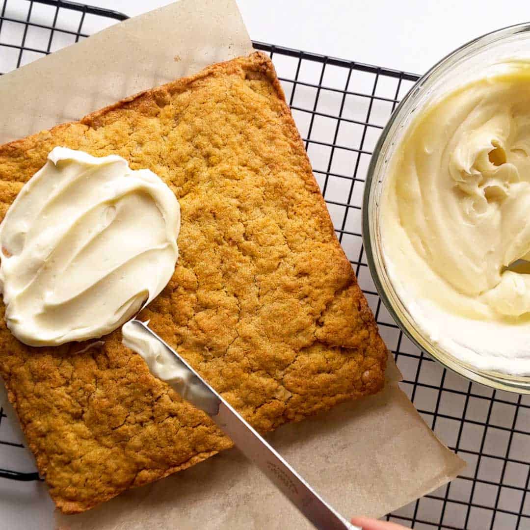 A knife spreads cream cheese frosting over the carrot cake tray bake.