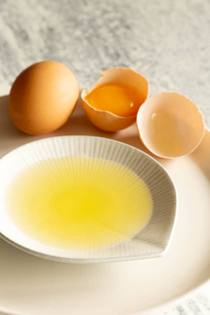 Egg white in a small dish, with an egg and egg shells in the background.