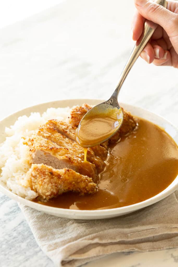A spoon drizzles curry sauce over the katsu.