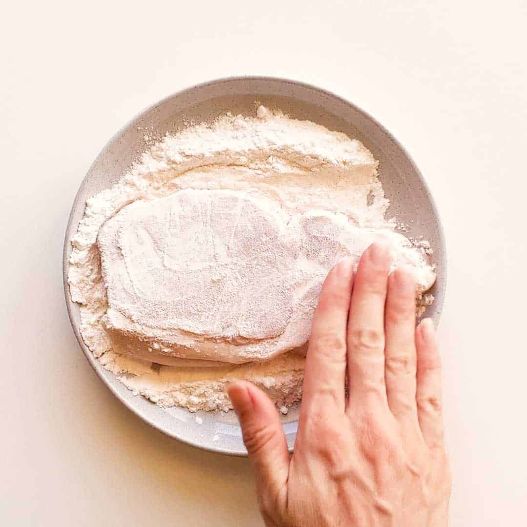 A hand pats flour over the pork cutlet.
