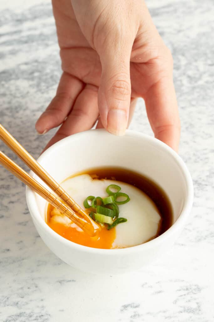 A hand holds a small bowl of Japanese hot spring eggs as the yolk is broken with chopsticks.