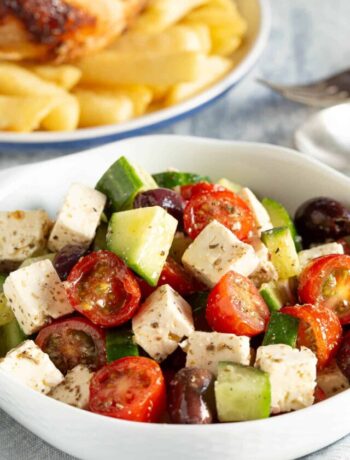 Greek salad in a white bowl with chicken and chips in the background.