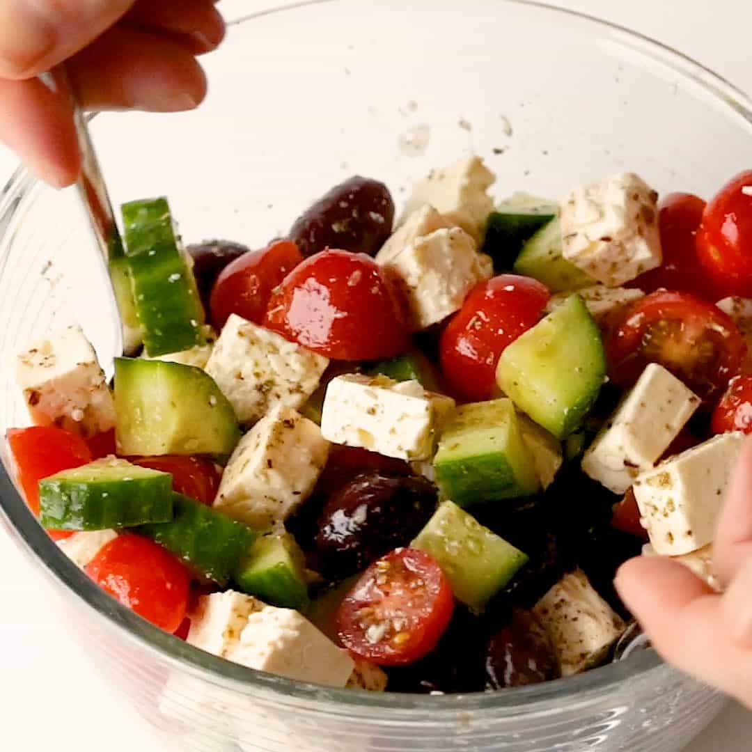 A Greek salad is tossed in a glass bowl using two spoons.