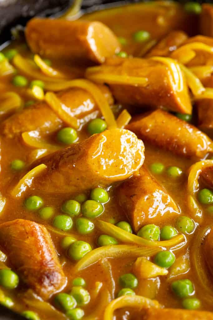 Close up of curried sausages in a pan showing the peas, onions and glossy curry sauce.
