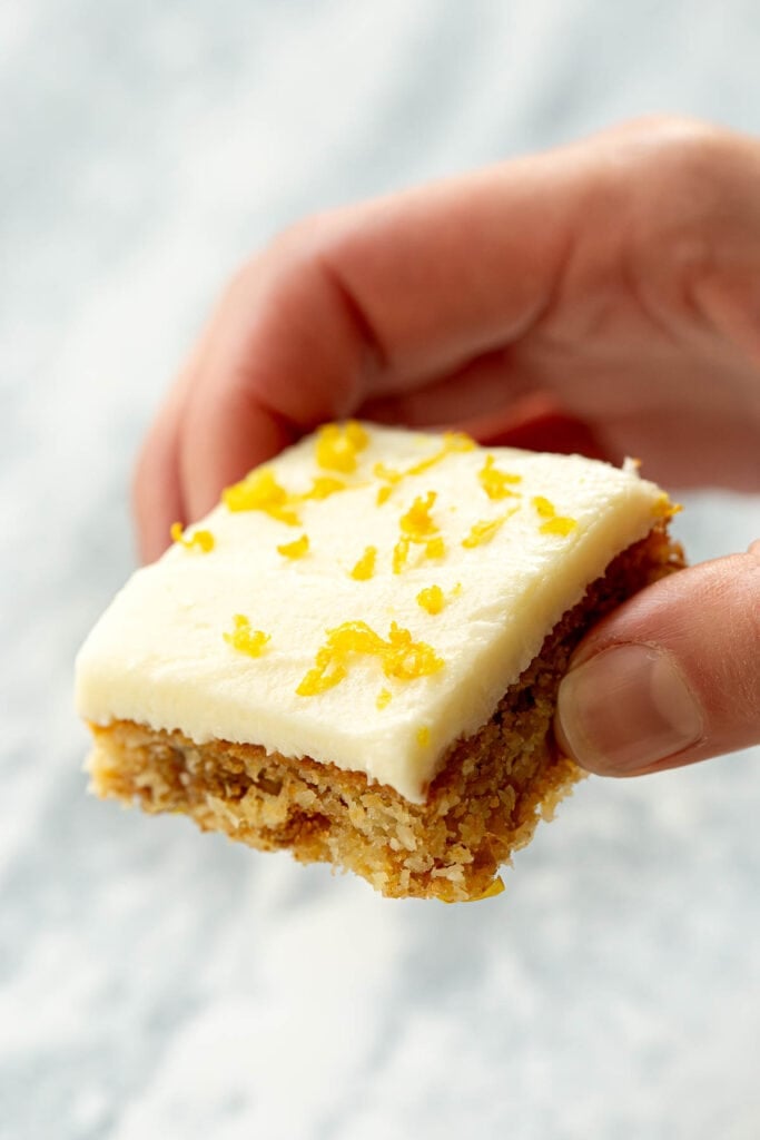 A hand holds a square piece of lemon Weetbix slice garnished with fresh lemon peel.