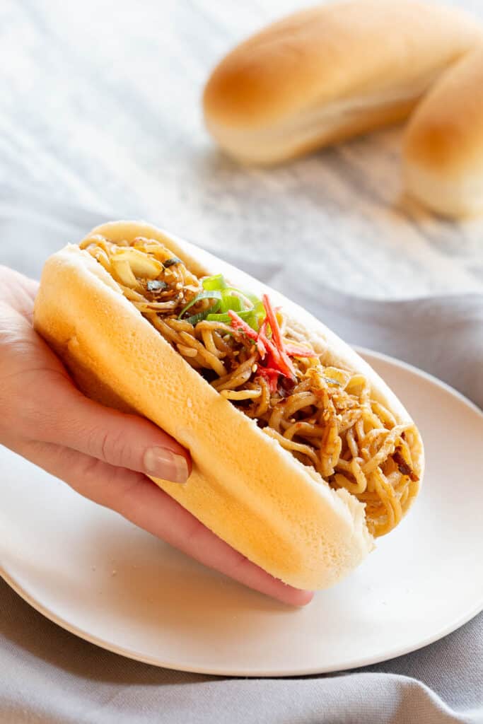 A hand holds a yakisoba pan over a white plate with hot dog buns in the background.