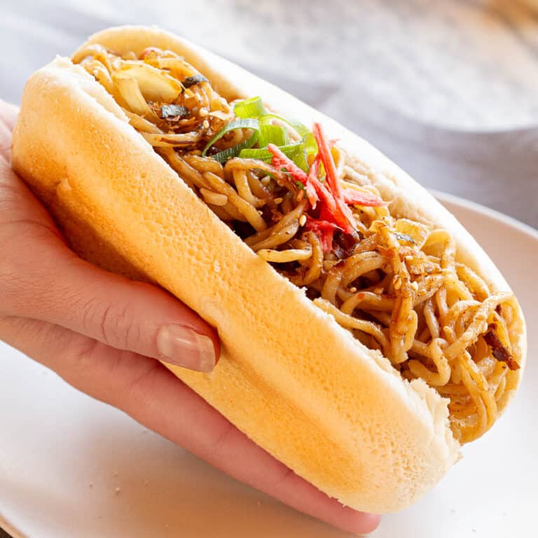 A hand holds a yakisoba pan garnished with pickled ginger and spring onion.