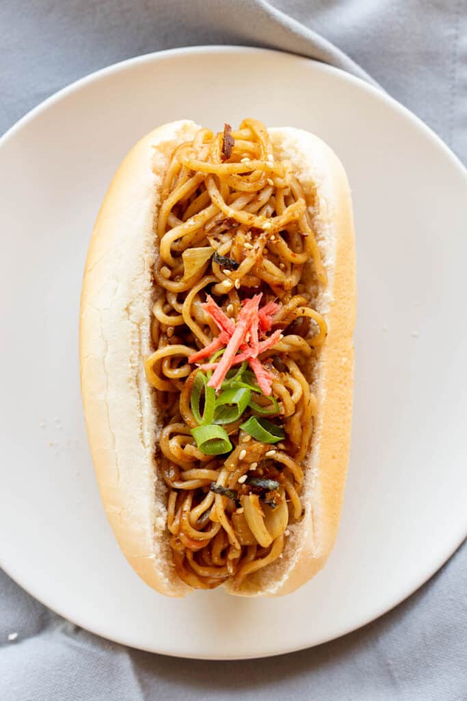 A hot dog bun filled with stir fried yakisoba noodles on a white plate.