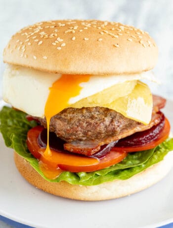 Burger on a plate loaded with egg, bacon, cheese, beetroot, tomato, lettuce and a chunky beef patty.