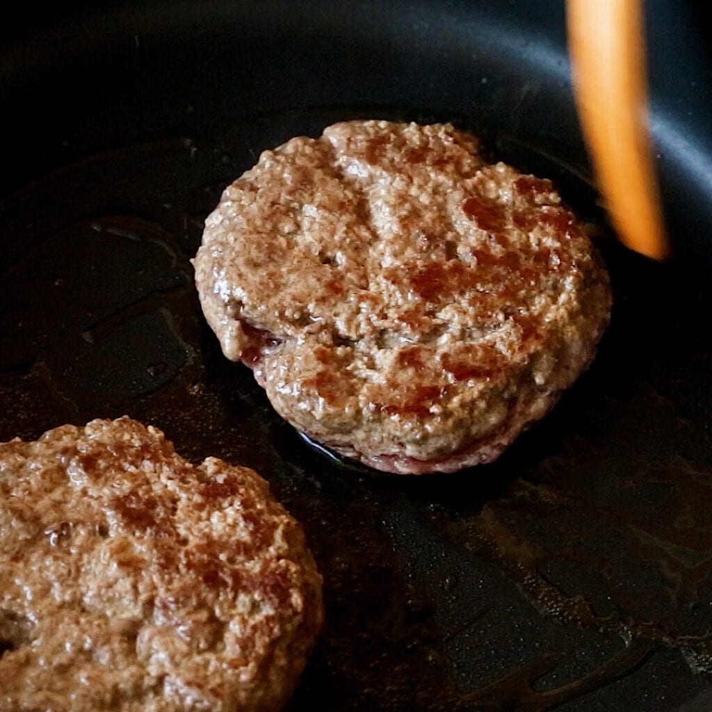 Beef patties on a frying pan, just flipped over with golden crust on top.