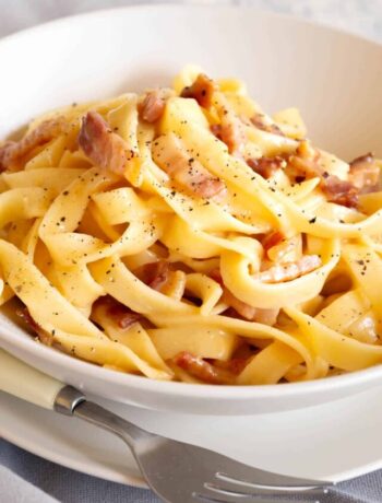 Ultra creamy and golden carbonara in a white bowl with a fork and plate.