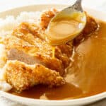 A spoon drizzling curry sauce over chicken katsu slices.