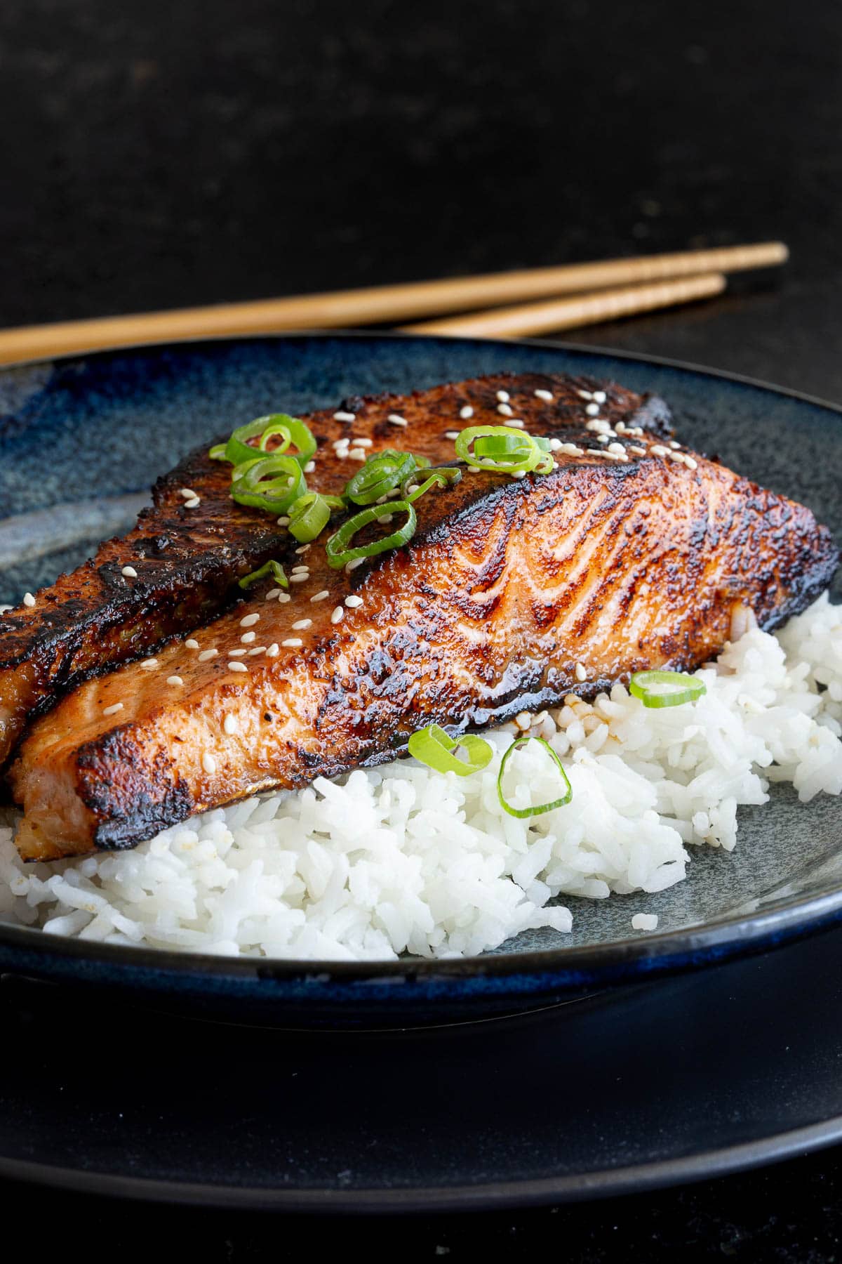 Two miso salmon steaks with a slight char on the side sit on a bed of white rice with chopsticks in the background.