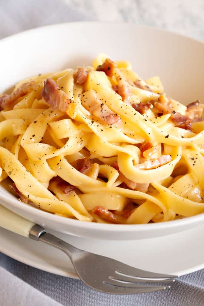 Ultra creamy and golden carbonara in a white bowl with a fork and plate.