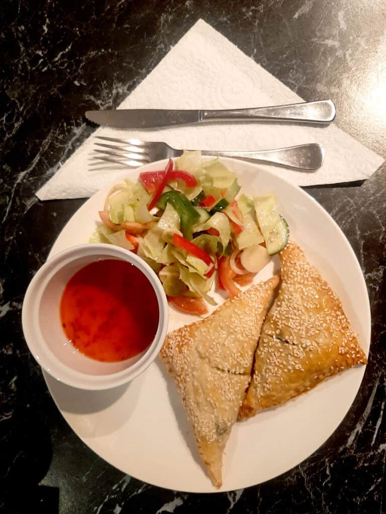A white plate of spinach and cheese triangles with Greek salad and sweet chilli sauce.