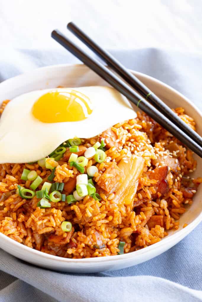 Kimchi fried rice in a white bowl with chopsticks, topped with egg, spring onion and sesame seeds.