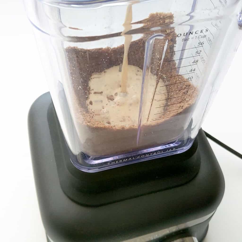 Pouring evaporated milk into the crushed spiced biscuits in a large blender.