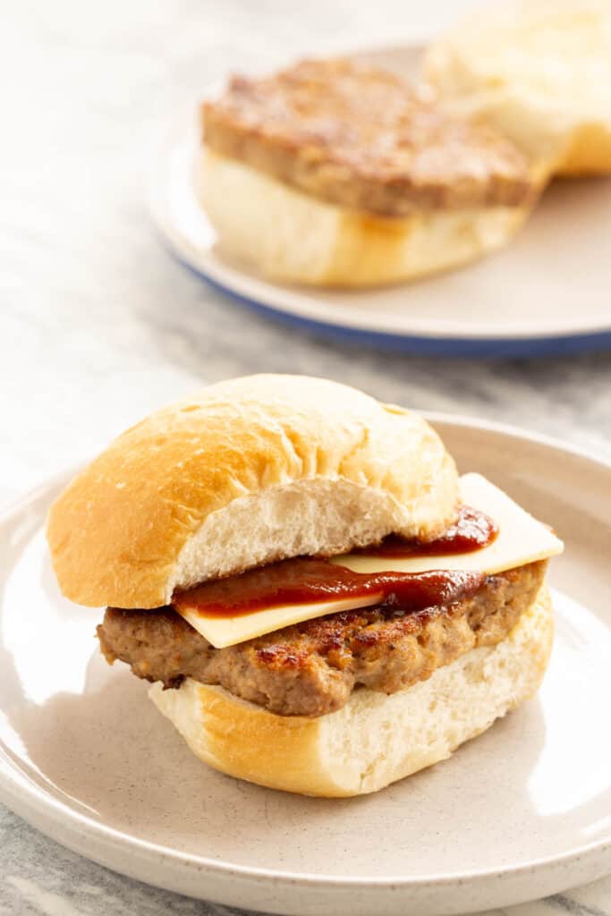 Two Scottish breakfast rolls stuffed with square sausage, cheese and brown sauce on white plates.