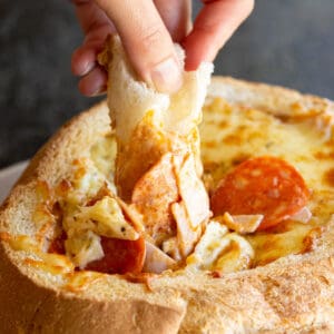 A piece of bread dips into the pizza dip with ham, pepperoni and cheese.