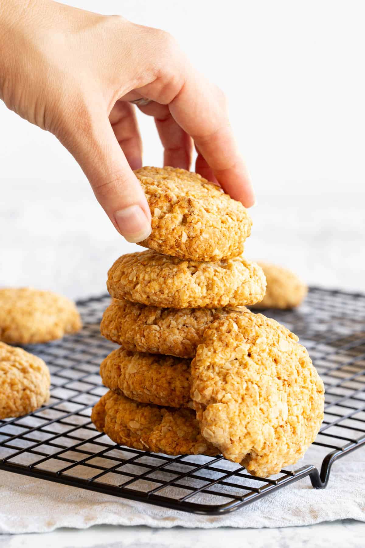 A stack of Anzac biscuits on a tray. A hand is placing a final biscuit on top.