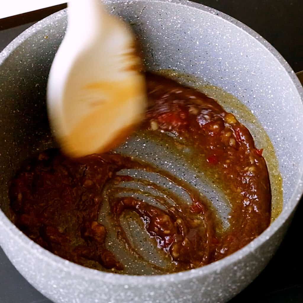 A white spoon stirring garlic, sambal oelek and tamarind puree in a small saucepan.