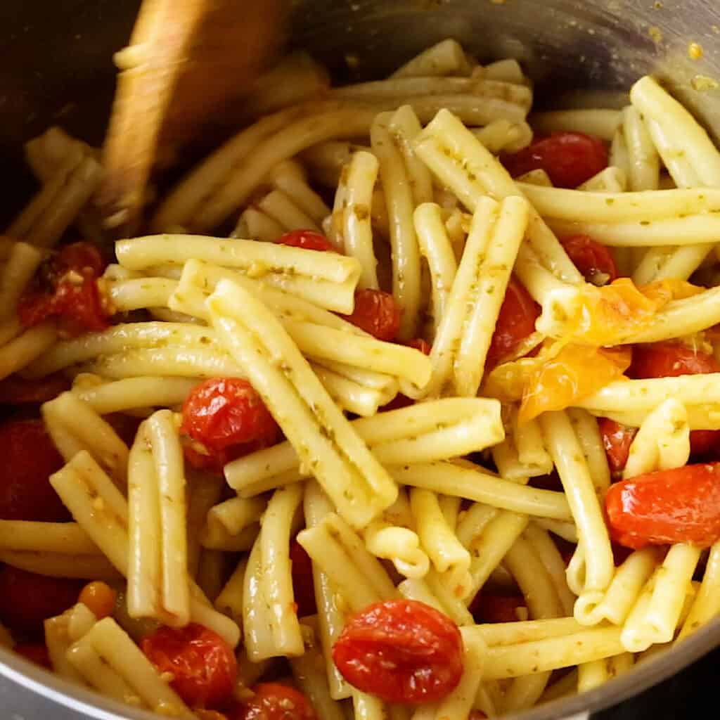 A wooden spoon mixes the roasted cherry tomato bursts through the pesto pasta.
